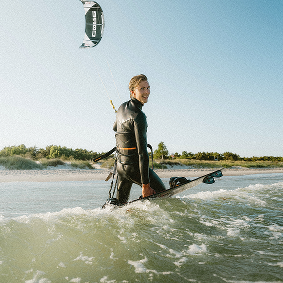 Person i våtdräkt som går i grunt vatten vid en strand och håller en kitesurfingbräda, med en kite i luften bakom sig.